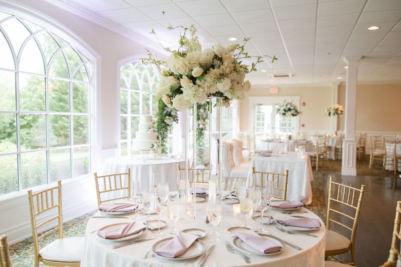 Reception table with tall white floral centerpiece beside arched windows